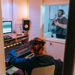 Músicos gravam uma canção em estúdio moderno enquanto um engenheiro de áudio opera a mesa de mixagem.