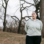 Mulher jovem com excesso de peso, usando roupa esportiva, se exercita em um parque arborizado.