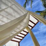 Pavilhão de madeira com cortinas brancas, palmeiras ao redor e céu azul com nuvens.