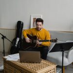 Homem de suéter amarelo toca violão em uma sala preparada para uma apresentação musical virtual.