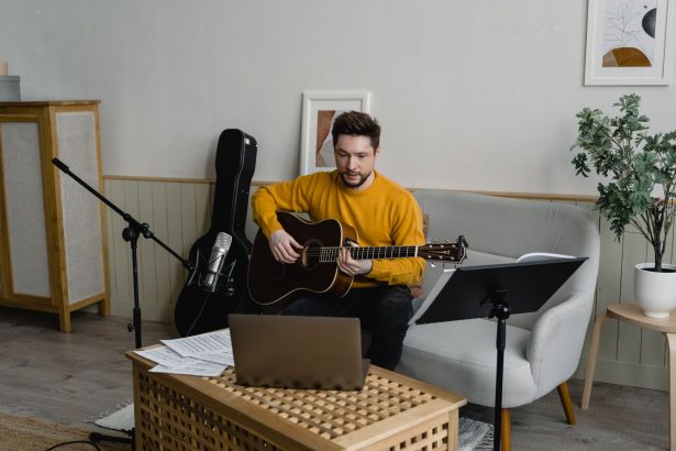 Homem de suéter amarelo toca violão em uma sala preparada para uma apresentação musical virtual.
